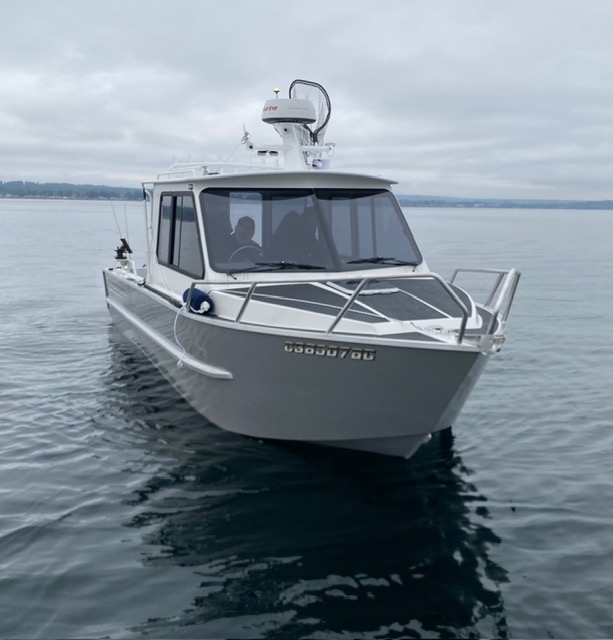 Kwigwis Adventures fishing charter boat viewed head-on from the water, displaying the hull and cabin with large front windows. The boat is equipped with radar and communication equipment mounted on top, set against a calm water backdrop with distant forested shoreline under overcast skies.